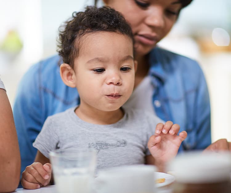 Niño pequeño sentado en la mesa sosteniendo una taza  y mamá sentada detrás de él